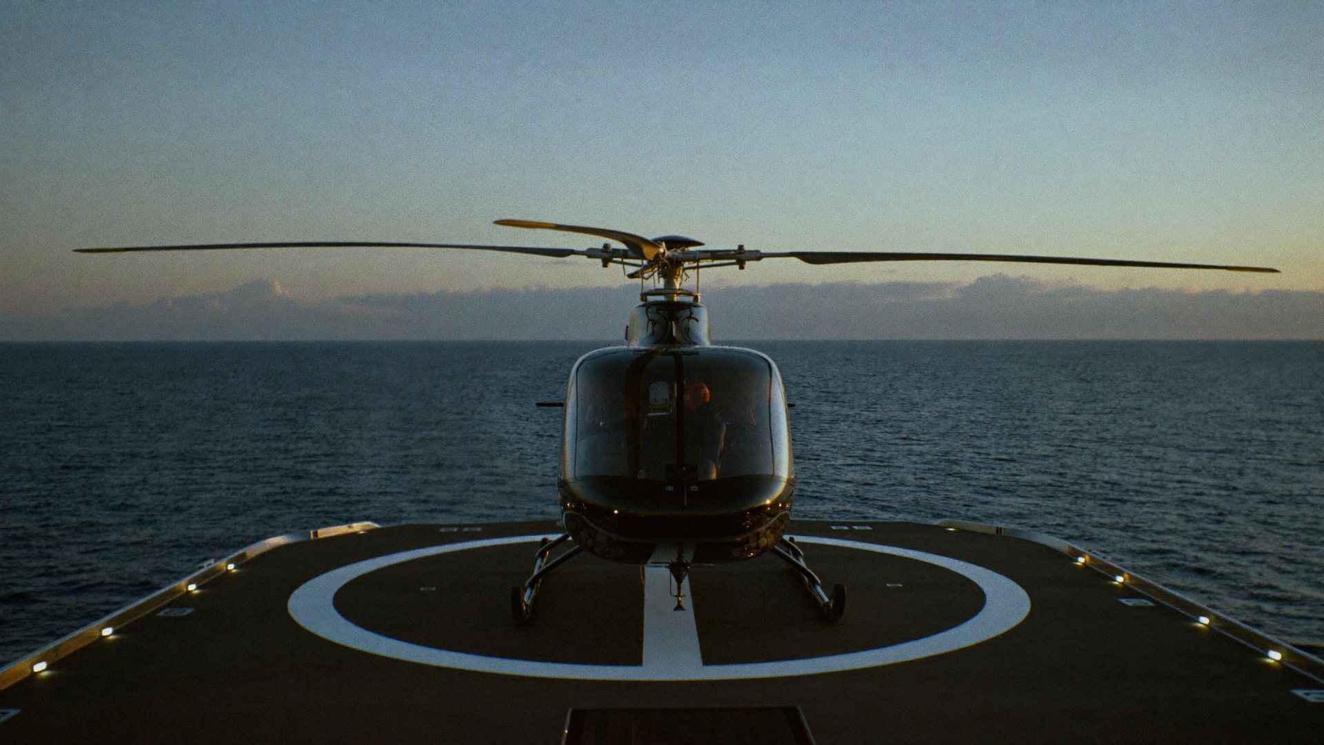 A helicopter at rest on a lit yacht helipad at blue hour, the open sea beyond.