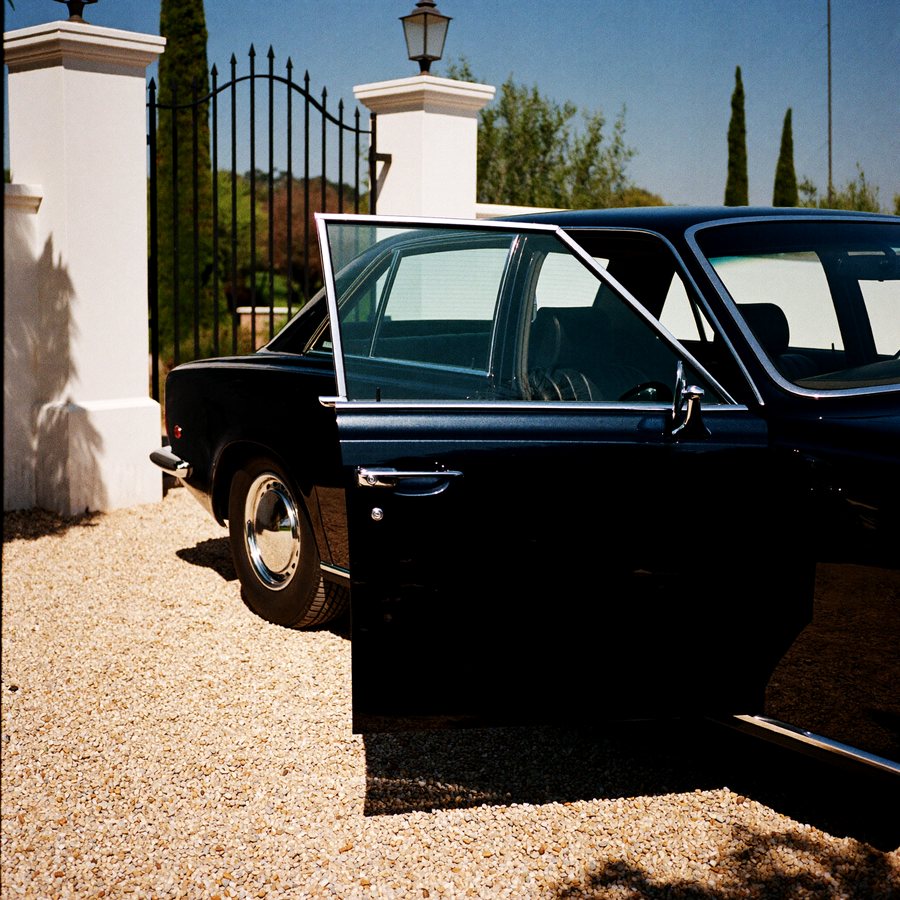 A dark saloon on pale gravel, door open, cypress shadow across it.