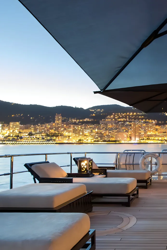 Monaco lit up at blue hour, seen across the water from a yacht's deck.