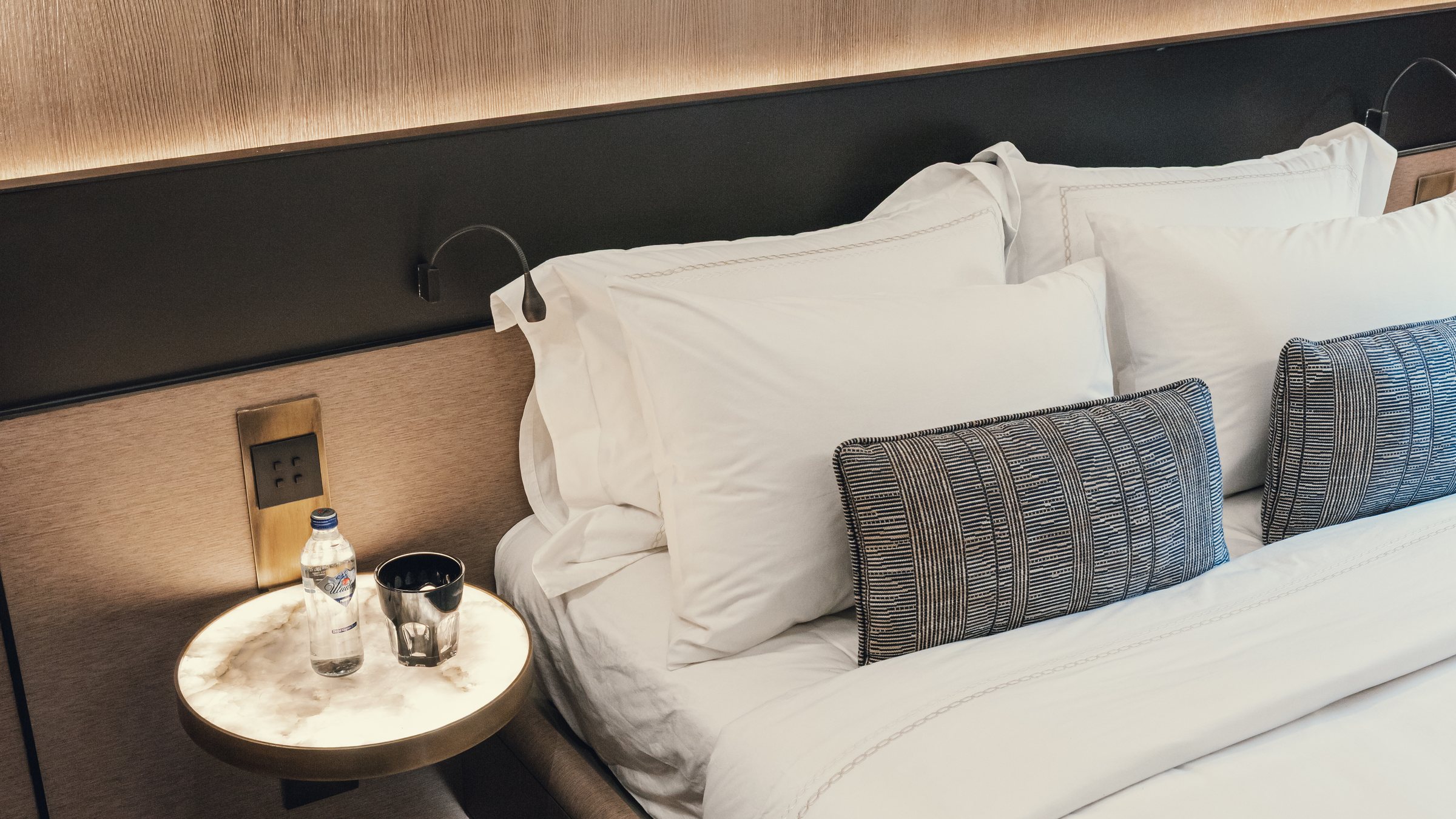 A made bed with white linen; cool blue and warm amber light drift slowly across it.